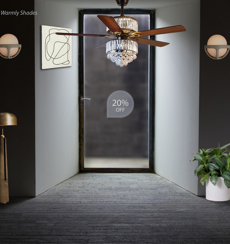 Puede incluir: Un ventilador de techo marr&oacute;n con detalles de cristal cuelga en una habitaci&oacute;n con una pared blanca y una puerta. El ventilador tiene aspas de madera y una l&aacute;mpara de ara&ntilde;a de cristal. Un cartel en la puerta dice "20% DE DESCUENTO".