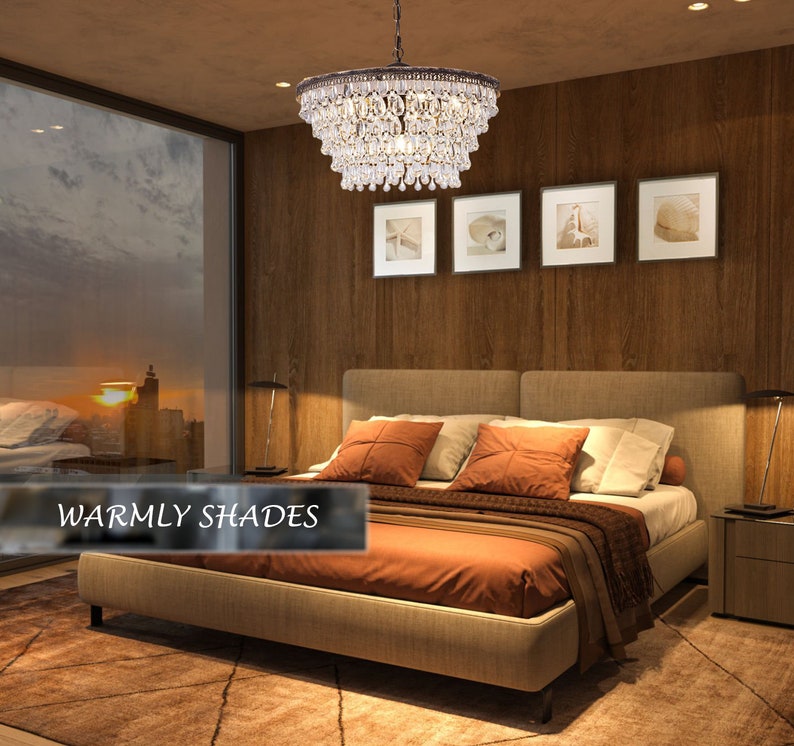 May include: A crystal chandelier with a brown metal frame hangs above a bed with a beige headboard and orange bedding. The room has a wooden wall and a window with a view of a city skyline at sunset. The text "Warmly Shades" is visible in the lower left corner of the image.