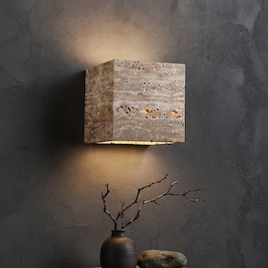 May include: A square wall sconce made of beige, porous stone with a natural, textured surface. The fixture emits a warm, soft glow from its top and bottom edges, casting light onto the wall above and below the rectangular stone housing.