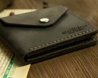 May include: A dark charcoal gray leather wallet featuring a triangular flap closure with a silver-toned metal snap button. The wallet has visible light-colored stitching along the edges and the brand name ecoWalnut wood-leather embossed on the front.