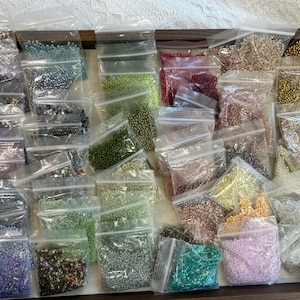 May include: A collection of small, clear plastic resealable bags filled with an assortment of tiny glass seed beads in various colors, including shades of blue, green, pink, yellow, white, metallic gold, and multi-colored mixes, arranged in a wooden tray.