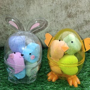 Könnte beinhalten: Zwei Osterbehälter gefüllt mit Marshmallow-Süßigkeiten. Einer ist ein durchsichtiger, hasenförmiger Behälter mit rosa und blauen Marshmallow-Hasen. Der andere ist ein gelber, kükenförmiger Behälter mit orangefarbenen und grünen Marshmallow-Küken.