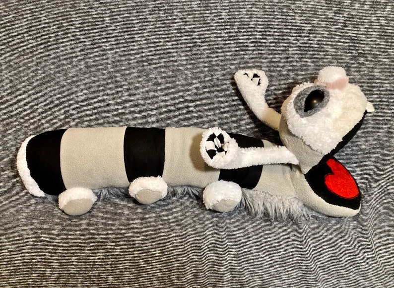 K&ouml;nnte beinhalten: Ein Pl&uuml;schtier in Form einer Raupe mit einem schwarz-grau gestreiften K&ouml;rper, wei&szlig;em Fell und einem roten Herzen auf dem Kopf.