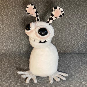 Peut inclure: Un jouet en peluche blanc avec de grands yeux noirs et un large sourire. Le jouet a des oreilles &agrave; carreaux noirs et blancs avec des accents roses. Le jouet a un corps blanc et moelleux et de longs bras et jambes blancs.