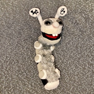 K&ouml;nnte beinhalten: Eine graue und wei&szlig;e Pl&uuml;schpuppe in Form einer Raupe mit einem gro&szlig;en, zahnigen Grinsen. Die Puppe hat gro&szlig;e, runde Ohren mit schwarz-wei&szlig; karierten Mustern.