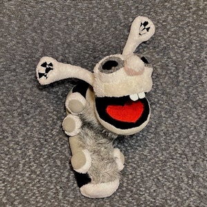 K&ouml;nnte beinhalten: Ein graues und wei&szlig;es Pl&uuml;schtier mit einem gro&szlig;en Mund und einem roten Herzen in der Mitte. Das Spielzeug hat einen langen, d&uuml;nnen K&ouml;rper mit mehreren kleinen, runden Gliedma&szlig;en.