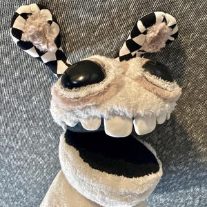 Peut inclure: Un jouet en peluche blanc avec des oreilles &agrave; carreaux noirs et blancs, de grands yeux noirs et une bouche grande ouverte avec des dents blanches. Le jouet est fait d'un tissu doux et duveteux et a un aspect fantaisiste et cartoon.