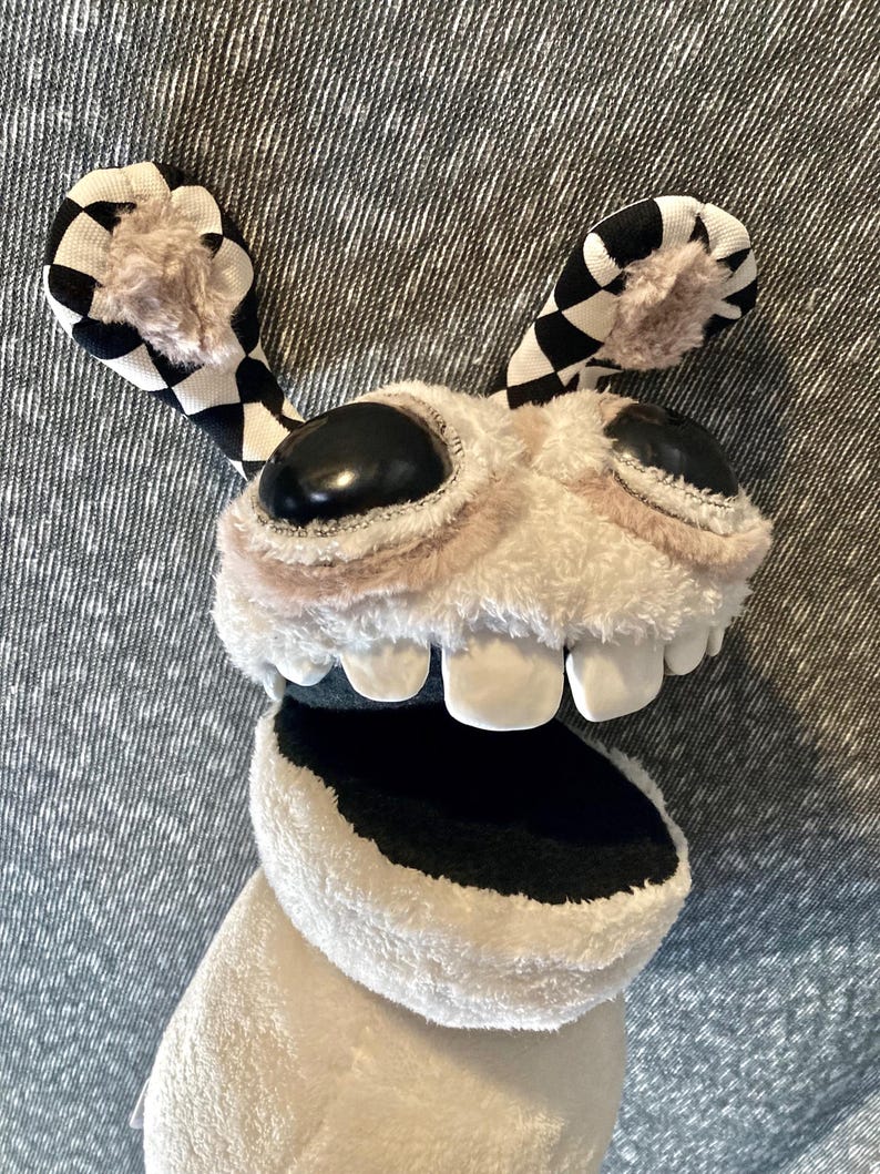 Peut inclure: Un jouet en peluche blanc avec des oreilles &agrave; carreaux noirs et blancs, de grands yeux noirs et une bouche grande ouverte avec des dents blanches. Le jouet est fait d'un tissu doux et duveteux et a un aspect fantaisiste et cartoon.