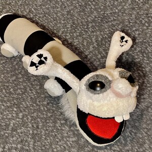 K&ouml;nnte beinhalten: Ein Pl&uuml;schtier in Form einer Raupe mit einem schwarz-wei&szlig; gestreiften K&ouml;rper und einem wei&szlig;en Kopf mit einem roten herzf&ouml;rmigen Mund. Das Spielzeug hat gro&szlig;e schwarze Augen und eine wei&szlig;e Nase.