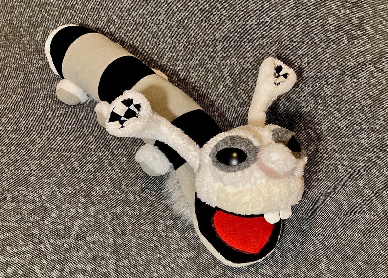 K&ouml;nnte beinhalten: Ein Pl&uuml;schtier in Form einer Raupe mit einem schwarz-wei&szlig; gestreiften K&ouml;rper und einem wei&szlig;en Kopf mit einem roten herzf&ouml;rmigen Mund. Das Spielzeug hat gro&szlig;e schwarze Augen und eine wei&szlig;e Nase.