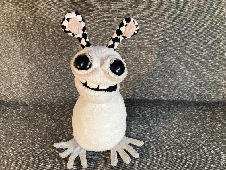 Peut inclure: Un jouet en peluche blanc avec de grands yeux noirs et un large sourire. Le jouet a des oreilles &agrave; carreaux noirs et blancs et de longs bras et jambes blancs et fins.