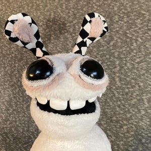 Peut inclure: Un jouet en peluche blanc avec de grands yeux noirs et un large sourire aux dents. Le jouet a des oreilles &agrave; carreaux noirs et blancs.