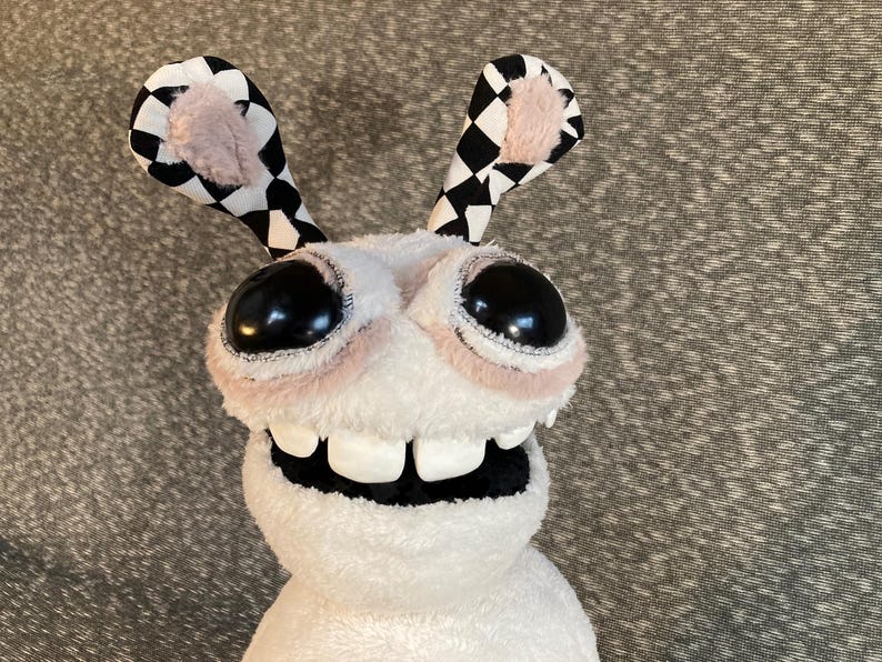 Peut inclure: Un jouet en peluche blanc avec de grands yeux noirs et un large sourire aux dents. Le jouet a des oreilles &agrave; carreaux noirs et blancs.