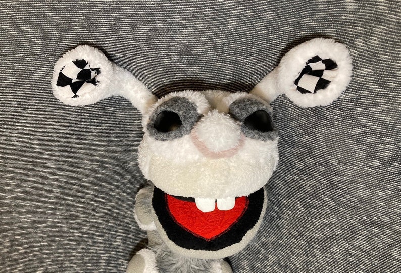 K&ouml;nnte beinhalten: Ein wei&szlig;es und graues Pl&uuml;schtier mit gro&szlig;en schwarzen Augen und einem roten Mund mit wei&szlig;en Z&auml;hnen. Das Spielzeug hat lange, schlaffe Ohren mit schwarz-wei&szlig;em kariertem Stoff auf der Innenseite.