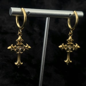 Peut inclure: Une paire de boucles d'oreilles créoles dorées avec des pendentifs en forme de croix ornées. Chaque croix présente une petite pierre précieuse transparente et des détails floraux complexes. Les boucles sont présentées sur fond sombre.
