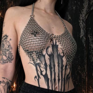 May include: A silver-toned chainmail halter top constructed from interlocking metal rings. The garment features a triangular silhouette with a neck strap and a central ring connector adorned with a small, dangling black cross charm.