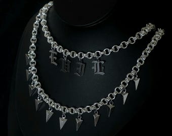 May include: A silver-toned necklace with two strands of chain. The top strand has the word "Evil" spelled out in gothic letters. The bottom strand has several triangular pendants. The necklace is displayed on a black mannequin.