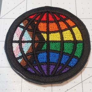 May include: A round embroidered patch with a globe design. The globe is made up of different colored stripes, including pink, blue, brown, green, yellow, orange, and red. The globe is surrounded by a black border.