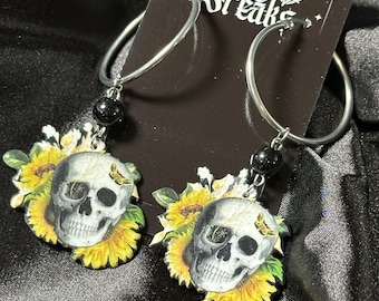 May include: A pair of silver hoop earrings with black beads and charms featuring a skull surrounded by sunflowers.