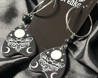 May include: A pair of black hoop earrings with a silver charm. The charm is a black and white image of a moth with a skull on its body, surrounded by crescent moons and stars. The charm is shaped like a triangle with a pointed top.