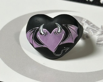 May include: A black heart-shaped ring with a purple devil heart design. The devil heart has white wings and horns.