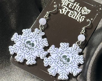 May include: A pair of silver snowflake earrings with a skull design. The snowflakes are white with black outlines and the skulls are black. The earrings are hanging from silver hooks with a light purple bead on each hook. The earrings are on a black background with the text "The Pretty Freaks" in white.
