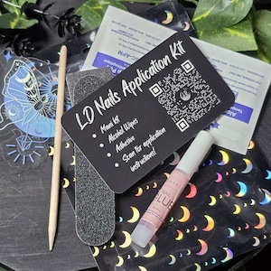 Könnte beinhalten: Eine schwarze Karte mit weißem Text, auf der "LD Nails Application Kit" steht und die Inhalte des Kits aufgelistet sind: Manikürset, Alkoholtupfer, Kleber und Scannen Sie die Anwendungshinweise. Die Karte hat einen QR-Code und befindet sich neben einer Flasche Nagelkleber, einer Nagelfeile und einem Holz-Nagelhaut-Schieber.