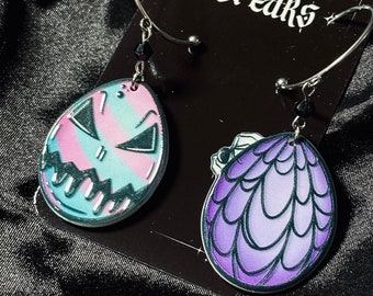 May include: A pair of earrings with silver hoops and dangling charms. One charm is a purple and white egg with a dragon design. The other charm is a pink and blue egg with a scary face.