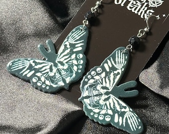 May include: A pair of dark gray butterfly earrings with a skeleton design. The wings feature a skull and bone pattern in white. The earrings have silver hooks and black bead accents. The tag reads "The Pretty Freaks".