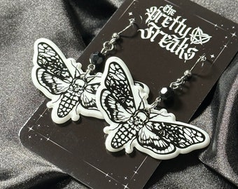 May include: A pair of black and white moth earrings with black bead accents. The earrings are on a card with the text "The Pretty Freaks" in a gothic font.
