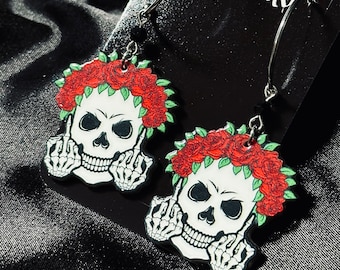 May include: A pair of skull-shaped earrings. Each earring features a white skull with a red rose crown and skeletal hands giving the middle finger. The earrings hang from silver-colored hooks with black beads.