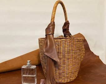 May include: A woven wicker basket bag with a curved handle and a brown leather tassel. A clear perfume bottle with the text "10 101 04" and a small lip gloss are next to the bag. The items are on a brown leather surface.