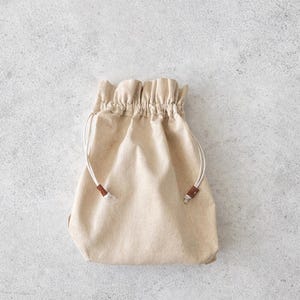 May include: A beige drawstring bag made of fabric. The bag has a gathered top and white drawstrings with brown leather accents. The bag is sitting on a light gray surface.