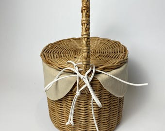 May include: A round, woven wicker basket with a lid and a handle. The basket is tan with a beige fabric lining and a white string tie closure. The handle is intricately woven.