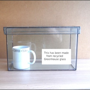 May include: A white mug with "COFFEE • TEA" printed on it, inside a clear glass display case. A label inside the case reads "This has been made from recycled Greenhouse glass."