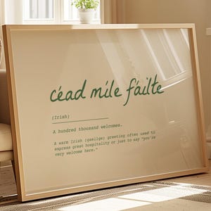 May include: A framed print with the words "céad mile fáilte" in green script. The text below explains the Irish greeting means "a hundred thousand welcomes." The print is in a light wood frame and leans against a sofa.