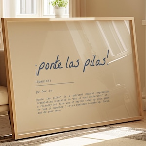 May include: A framed print featuring the Spanish phrase "¡ponte las pilas!" in blue script, with the English translation "go for it." and a definition. The light wood frame complements the print, displayed in a living room setting.