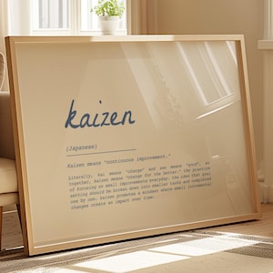 May include: A framed print with the word "kaizen" in blue script. The print includes the definition of "kaizen" in smaller text below the word. The frame is a light wood color, and the print is displayed in a well-lit room.