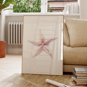 Puede incluir: Una impresión enmarcada con una estrella de mar en acuarela en tonos rosas y morados. La obra de arte se exhibe en un salón luminoso con un sofá beige, una silla de madera y una planta en maceta. Un póster enrollado y libros están sobre la alfombra.