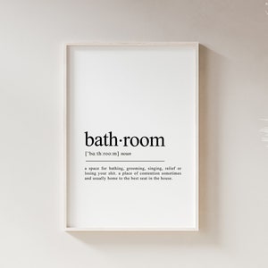 May include: A black and white printable wall art print with the word "bath-room" defined as a space for bathing, grooming, singing, relief or losing your shirt, a place of contention sometimes and usually home to the best seat in the house. The print is framed in a white frame and is hanging on a white wall. A white pampas grass bouquet is in a woven vase on a white shelf.