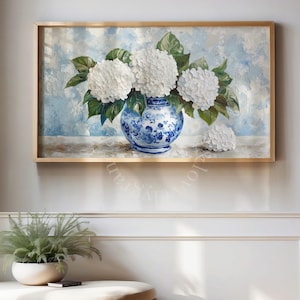 Pode incluir: Uma pintura emoldurada de hortênsias brancas em um vaso azul e branco. O vaso está sobre um fundo azul claro e branco. A pintura está em uma moldura dourada e está pendurada em uma parede branca.