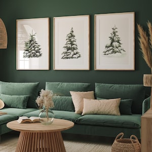 May include: A green velvet sectional sofa with a light brown throw blanket, two pillows, and a coffee table with a book on top. Three framed illustrations of snow-covered pine trees hang on the wall behind the sofa.