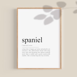 Puede incluir: Una impresión de arte de pared imprimible en blanco y negro con la palabra "spaniel" definida como una raza de perro feliz, juguetona y cariñosa con pelo largo y ondulado y orejas caídas. La impresión está enmarcada en un marco de madera marrón claro.