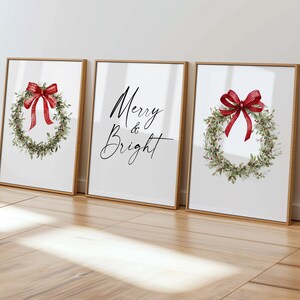 May include: Three framed art prints with a Christmas theme. Two prints feature a watercolor wreath with a red bow. The center print has the words "Merry & Bright" in elegant script. The frames are a light brown color.