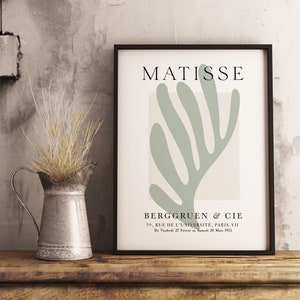 Op de afbeelding: Een zwart ingelijste print met een lichtgroen abstract bladontwerp en de tekst "MATISSE" en "BERGGRUEN & CIE" in wit. De tekst onder het ontwerp luidt "70. RUE DE L'UNIVERSIT&Eacute;, PARIS-VII" en "Du Vendredi 27 F&eacute;vrier Samedi 28 Mars 1953".
