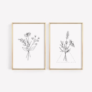 May include: Two black and white line drawings of floral bouquets in gold frames. The first bouquet features a daisy and other wildflowers. The second bouquet features a daisy, lavender, and other wildflowers.