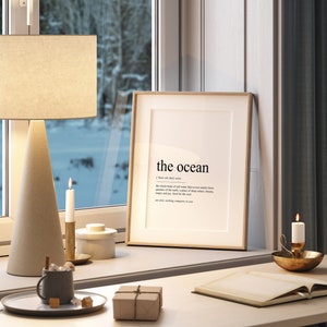 May include: A framed print with the word "the ocean" and its definition. The print is on a white background with black text. The print is sitting on a white table with a lamp, candles, and a book.