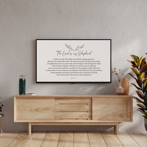 Puede incluir: Un mueble de TV de madera con un televisor de pantalla plana que muestra una impresi&oacute;n en blanco y negro del Salmo 23, "El Se&ntilde;or es mi pastor". El televisor est&aacute; en un sal&oacute;n con una planta en maceta, una l&aacute;mpara de pie y una mesita auxiliar.