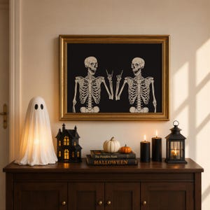 May include: A framed wall art piece featuring two skeletons on a black background, one making a rock-and-roll hand gesture. The display includes a white ghost lamp, a black house lantern, two pumpkins, two black candles, and two books.
