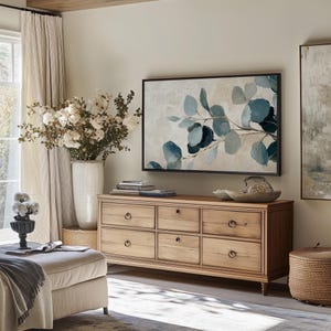May include: A light wood dresser with six drawers and brass knobs. The dresser is in a living room with a large TV above it, a vase of white flowers, and a throw blanket on a light-colored ottoman.